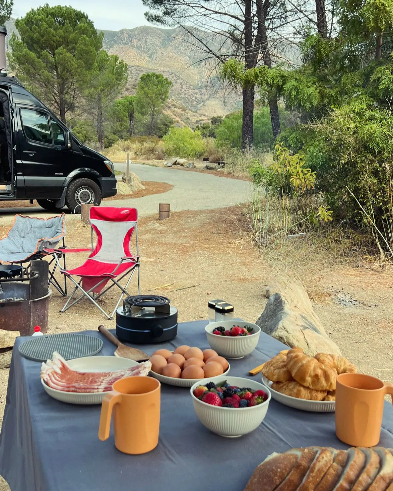 A camp breakfast of bacon, eggs, berries, and croissants set out around ElementOne, with a camper van parked below the mountains